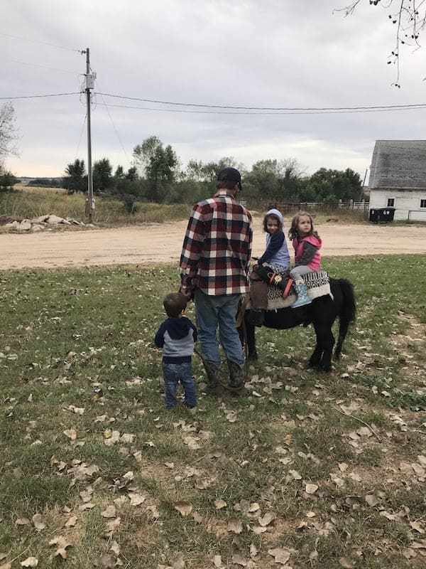 dad, 3 little children, and a mini horse on a family ranch with a white barn in the background