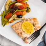 white plate with the cooked salmon and a lemon slice on top, and cooked veggies to the side.
