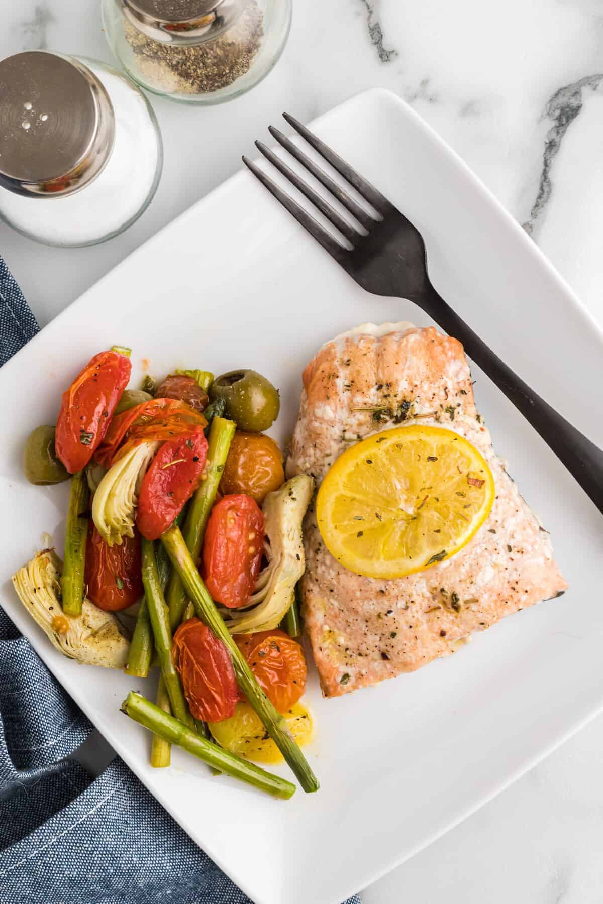 square white plate with the cooked salmon recipe and cooked asparagus, artichokes, olives and tomatoes to the side.