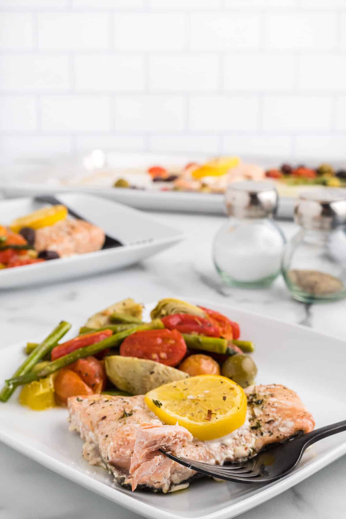 square white plate with the cooked salmon recipe and cooked asparagus, artichokes, olives and tomatoes to the side, and a forkful of flaky salmon.