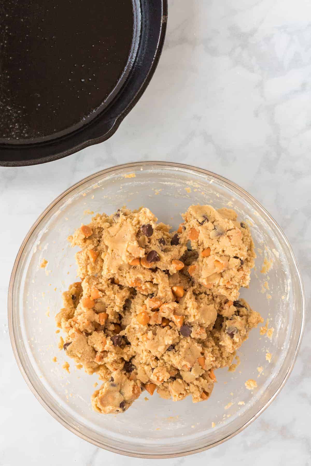 the cookie dough in a large glass bowl with the cast iron skillet in the background