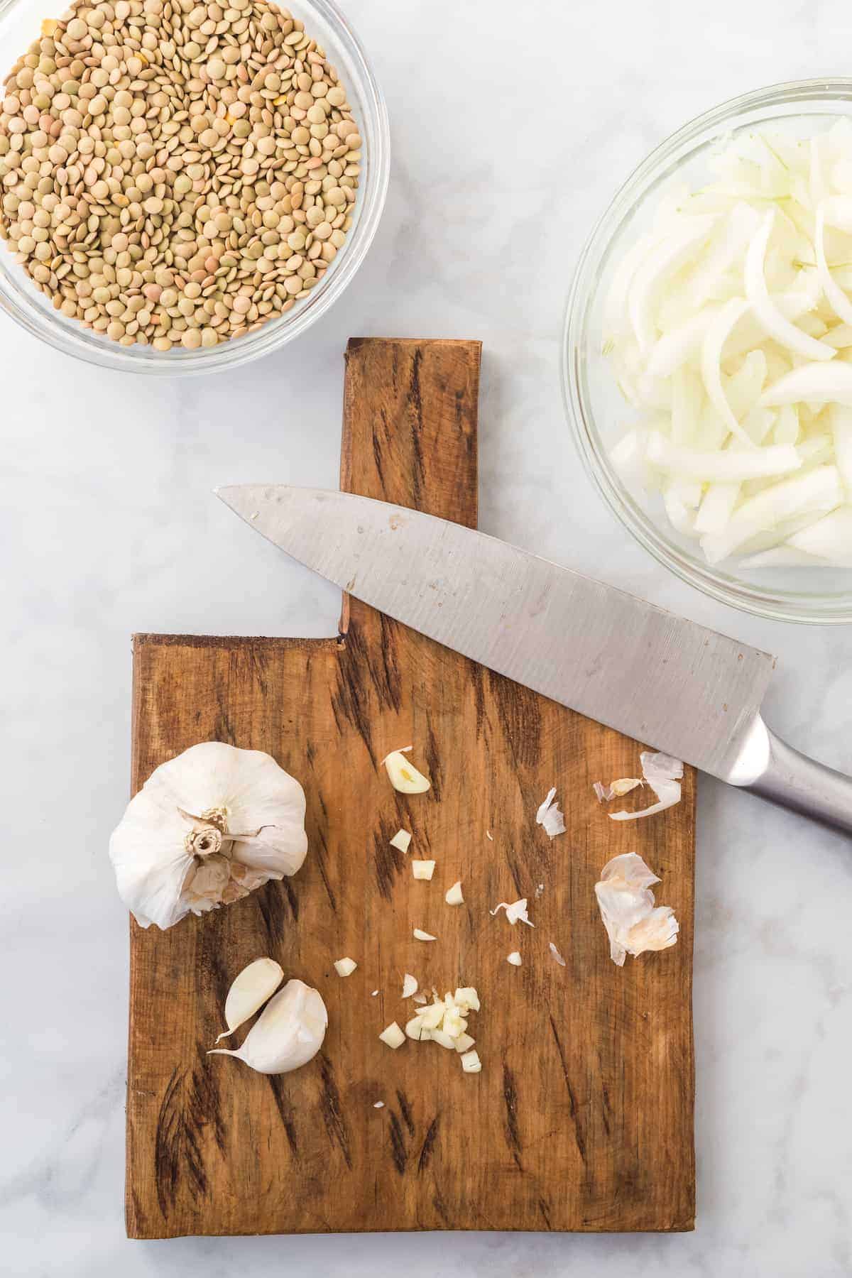 chopping the garlic on a cutting board with the onions and lentils in bowls in the background