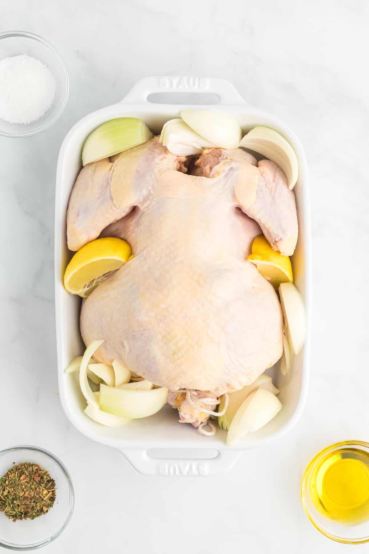 lemon wedges and onion quarters spread around the whole chicken in a white baking dish