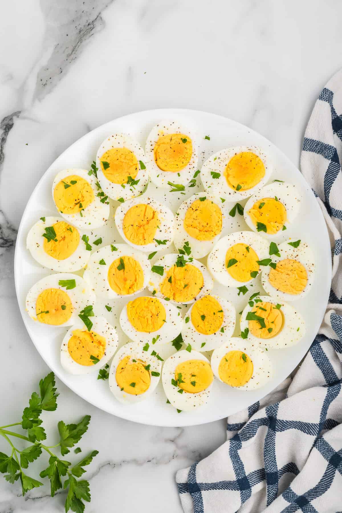 Ninja Foodi hard boiled eggs sliced, on a white platter with chopped herbs an fresh cracked black pepper over top. And a black and white checked tea towel rests to the side. 