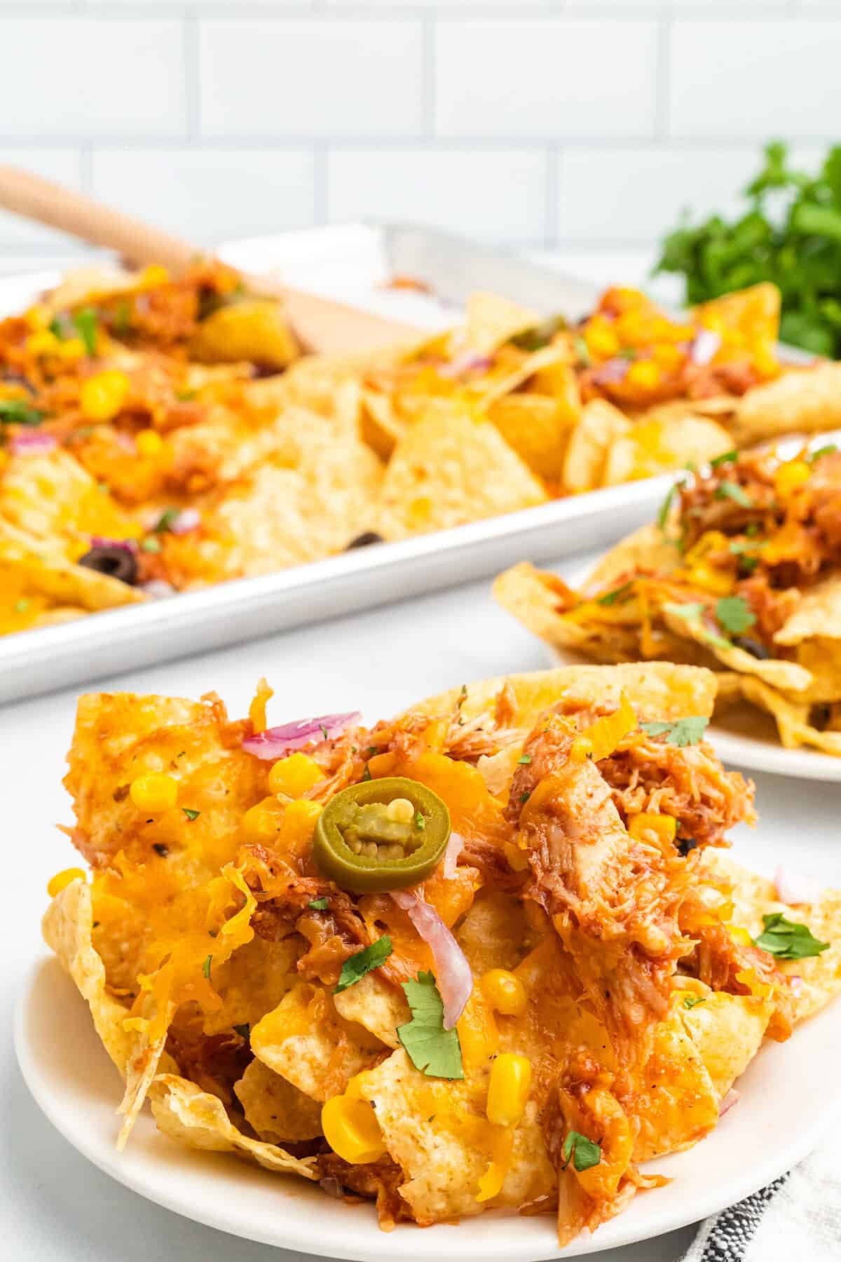 sheet pan of bbq chicken nachos with a white plate in front of it loaded with toppings.