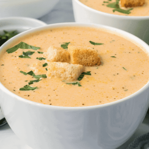 closeup of white bowl filled with creamy acorn squash soup with croutons and fresh herbs on top