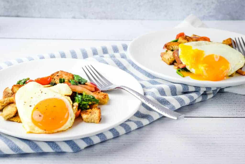 two plates of breakfast panzanella on a table with forks and blue striped tea towel