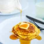 white plate with a stack of fresh pancakes, butter and pouring maple syrup from a small pitcher
