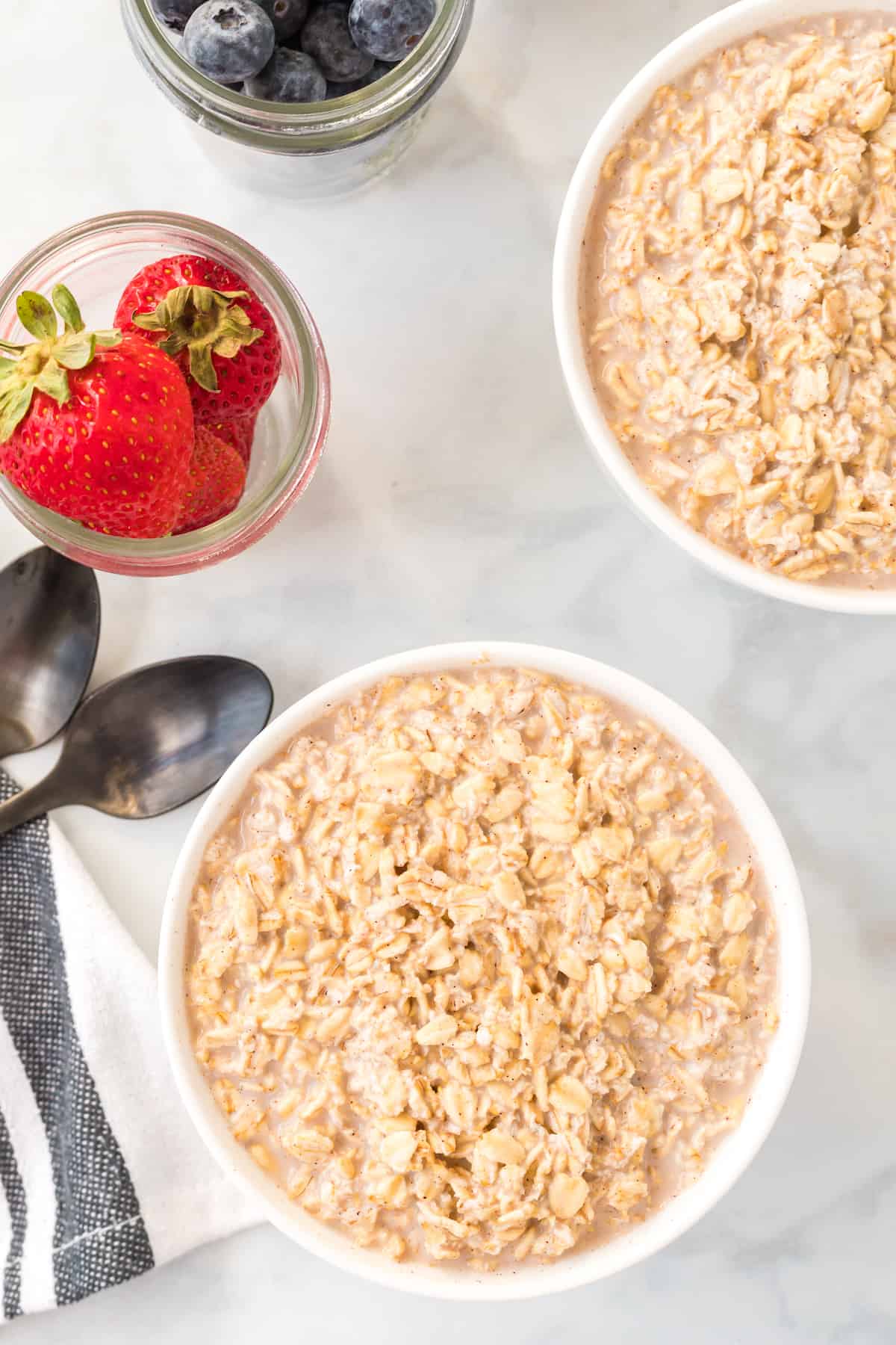 two white bowls of overnight oats with jars of fresh strawberries and two black spoons besides the bowls