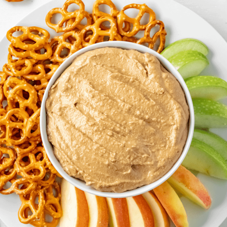 white platter with a white bowl in the center with the gingerbread hummus and apple slices wrapping around the front and right side. Pretzels wrapping around left and back side.