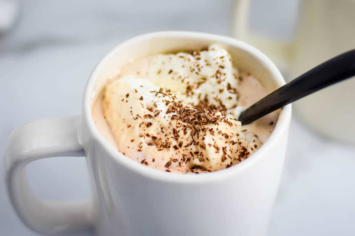 maple whipped cream topped on homemade hot cocoa in a white much with a black spoon inside