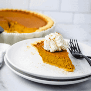 two white plates stacked with a slice of pumpkin pie and homemade whipped cream on top. the pan of pumpkin pie is in the background