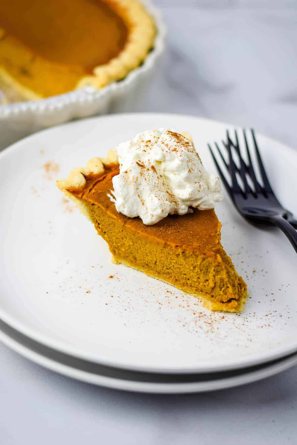 homemade whipped cream on top of the pumpkin pie. the pie is on top of a stack of white plates with 2 black forks resting beside it.