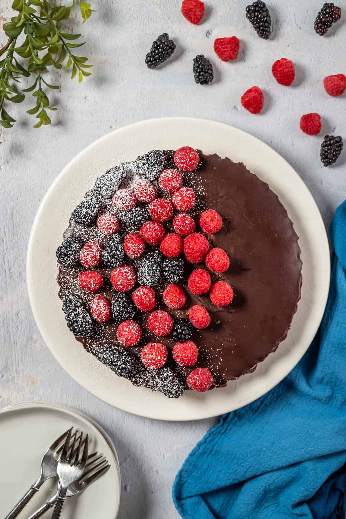 white plate of chocolate tart with fresh berries on top and stack of white plates with forks resting beside it 
