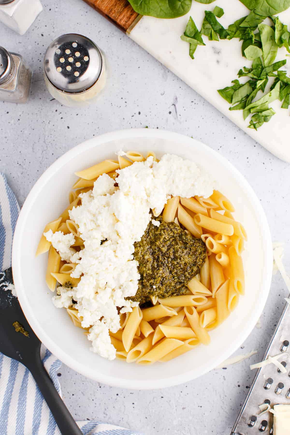 pesto and ricotta cheese added to the top of the pasta in the large white bowl