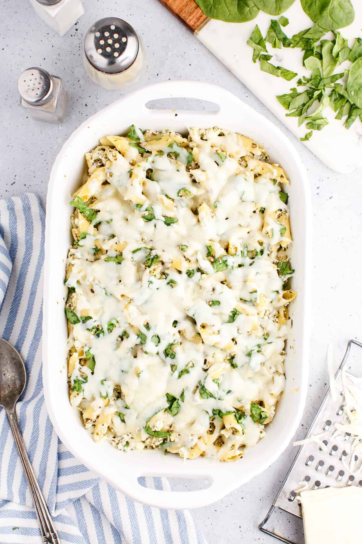 white casserole dish with baked pesto pasta and blue and white striped tea towel to the side with a serving spoon to the side and chopped basil on a cutting board