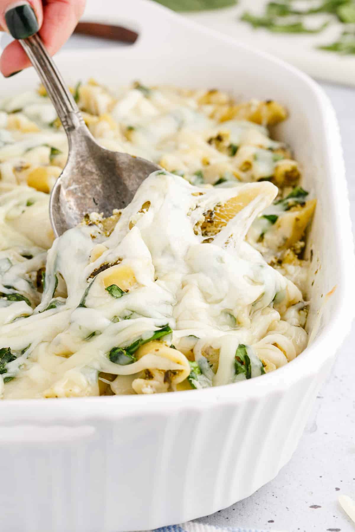 white casserole dish with pesto pasta and handing a serving spoon