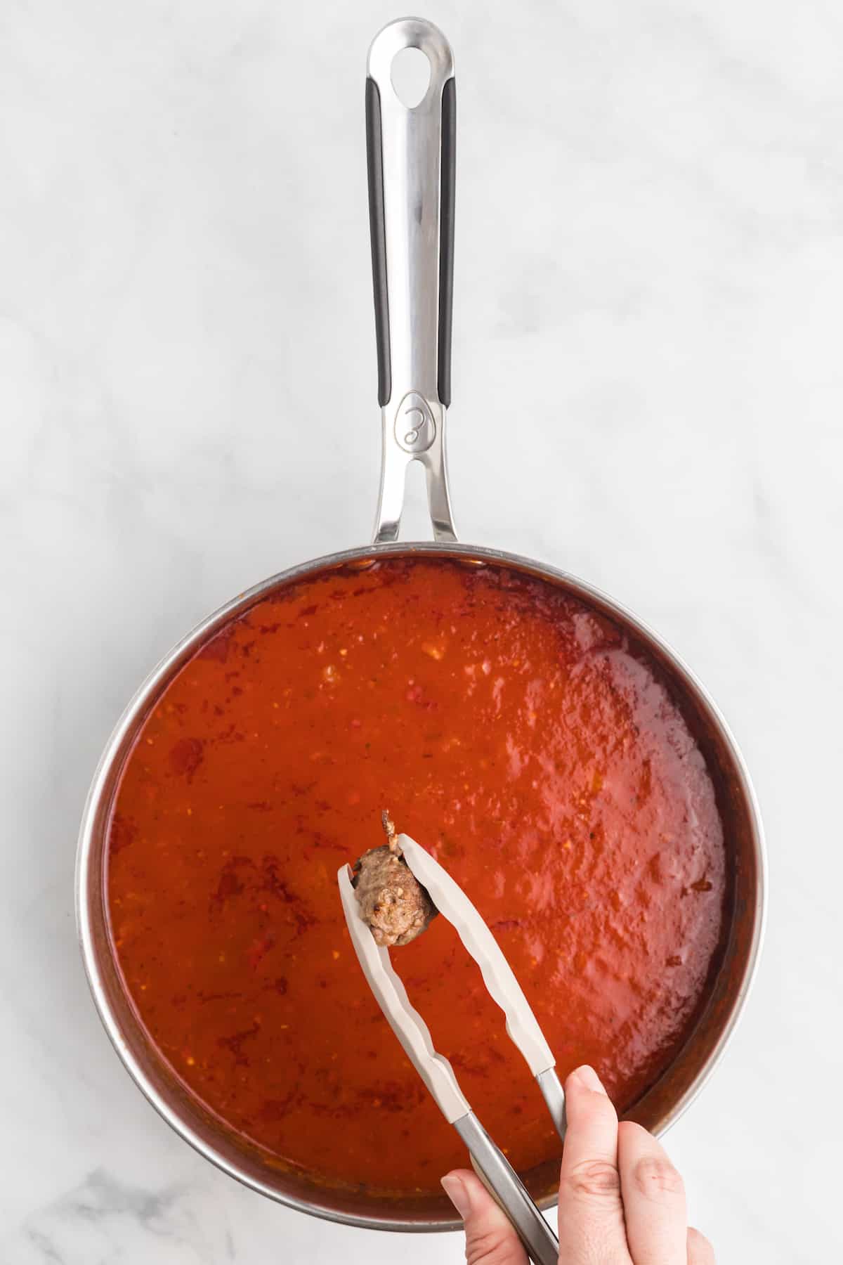 using tongs to add the cooked meatballs to the marinara sauce.