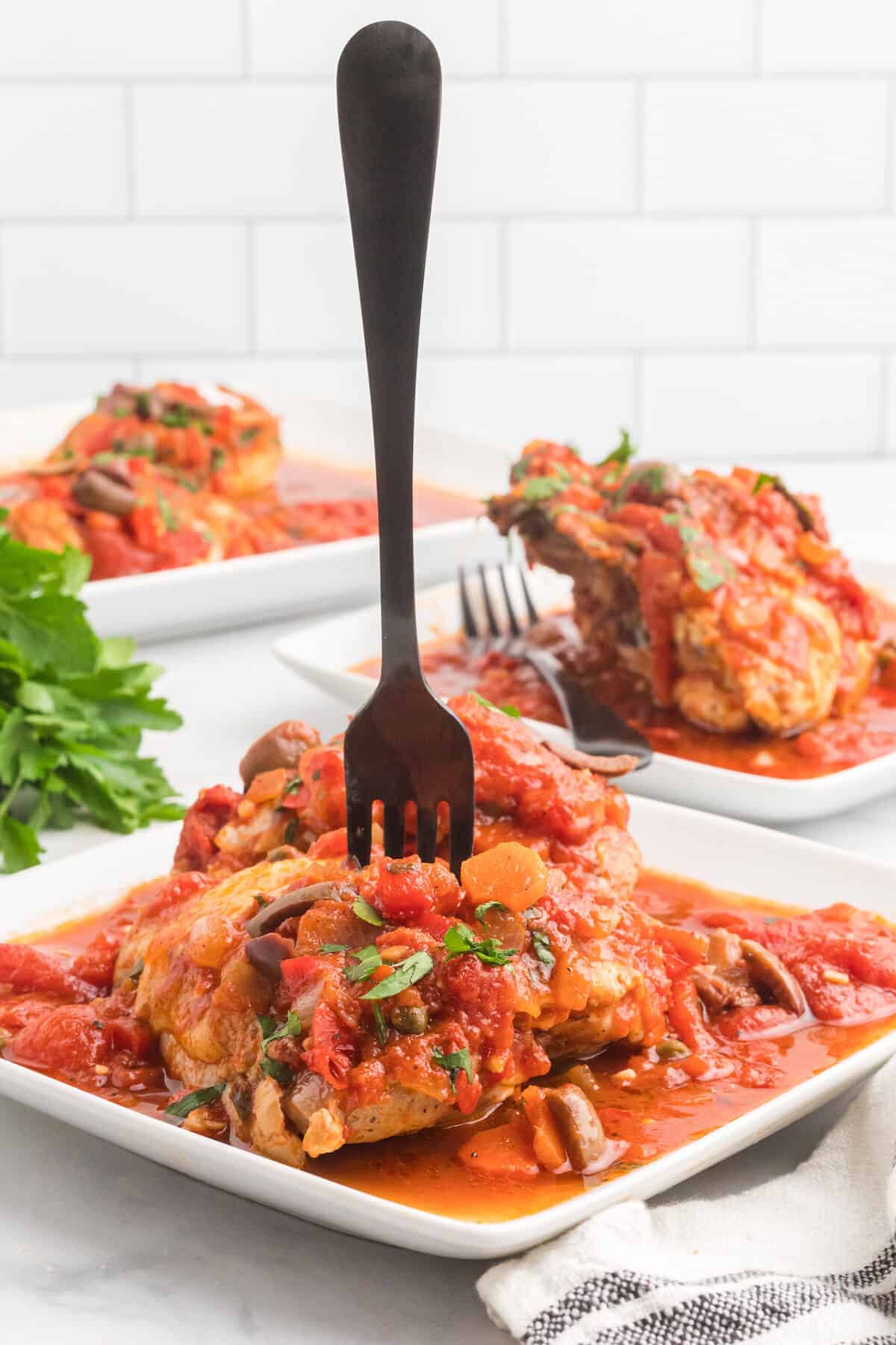 two white square plates of chicken cacciatore with a black fork sticking out of one of the chicken breasts.
