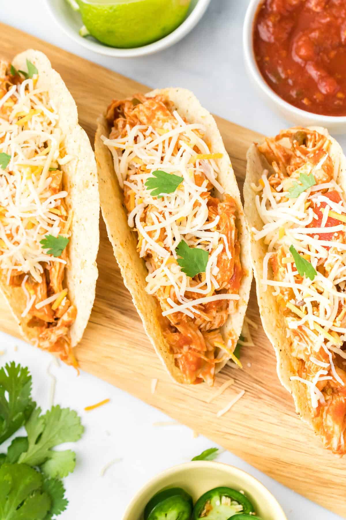 wooden board with shredded chicken tacos and small bowls of garnishes.