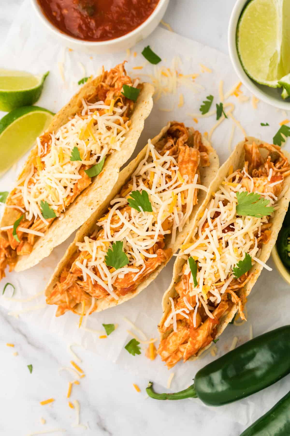three shredded chicken tacos with cheese, cilantro, jalapeño and salsa garnishes.