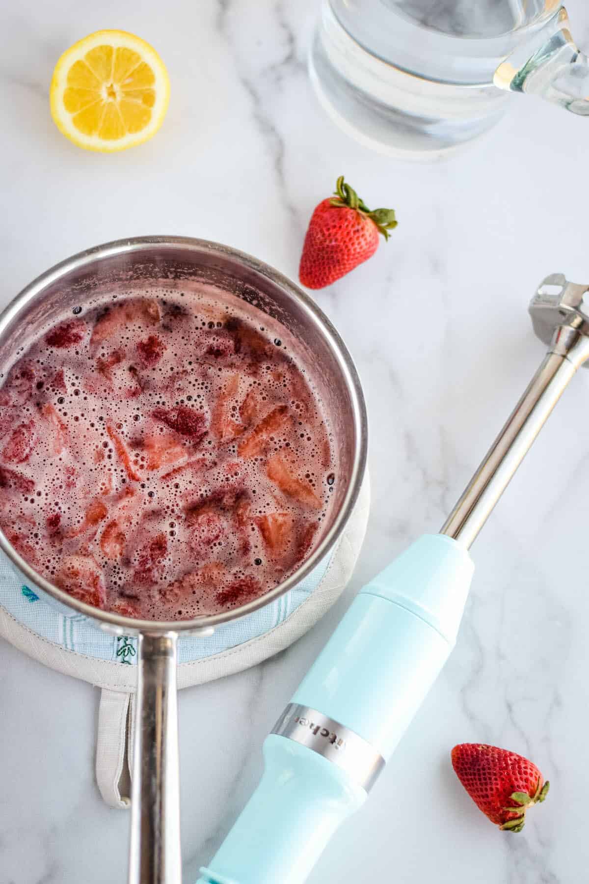 Using a blue stick blender to mix the strawberries with honey and water in a saucepan. 