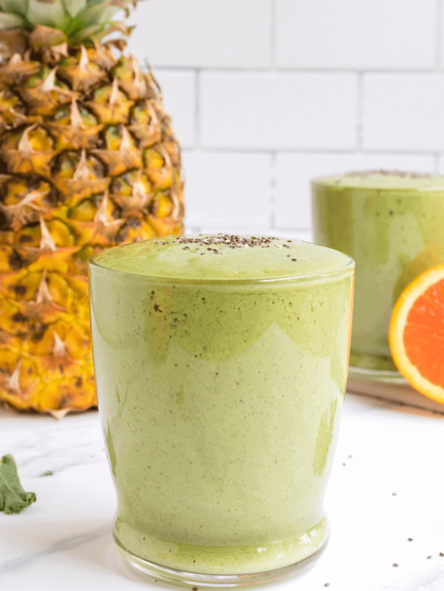two glasses of the tropical green smoothie with a pineapple and orange in the background.