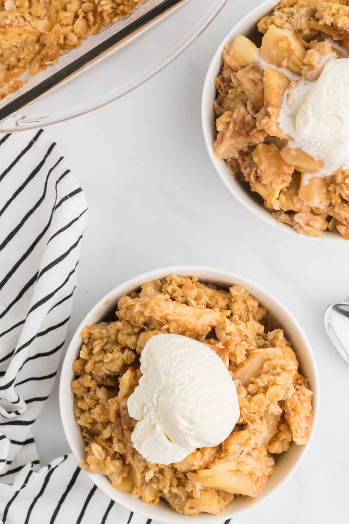 two white bowls of apple crisp with a scoop of vanilla ice cream on top.