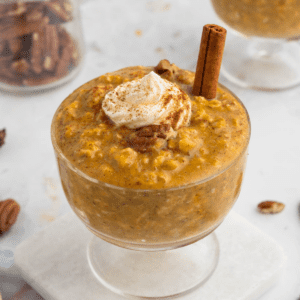 glass bowl with pumpkin overnight oats, whipped cream on top, and cinnamon stick to the side.
