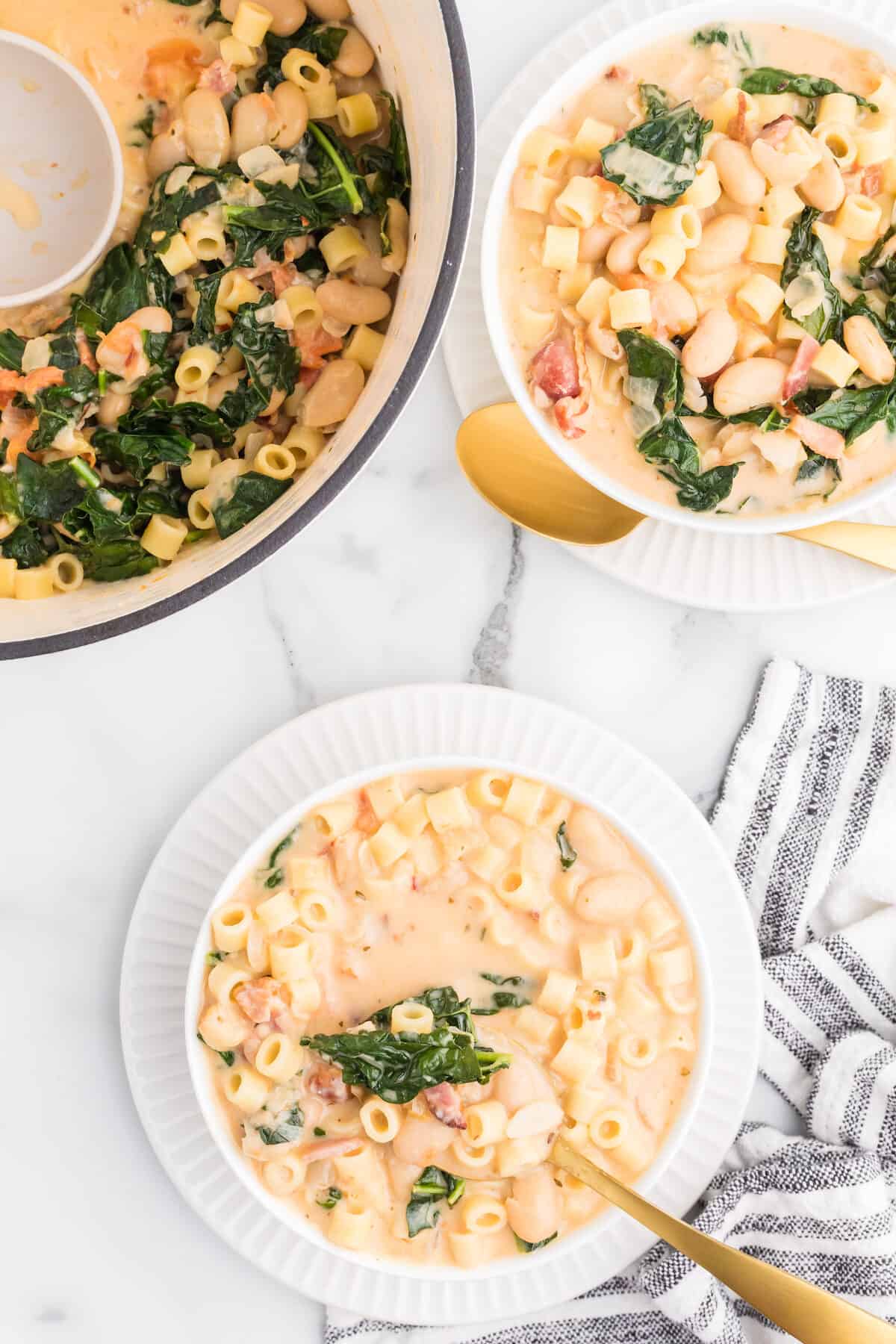 two white bowls of creamy pasta fagioli soup with the dutch oven of soup.