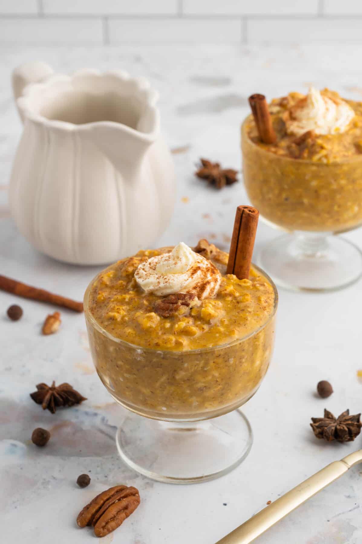two glass bowls with pumpkin pie oats and a small white pitcher with maple syrup to the back.