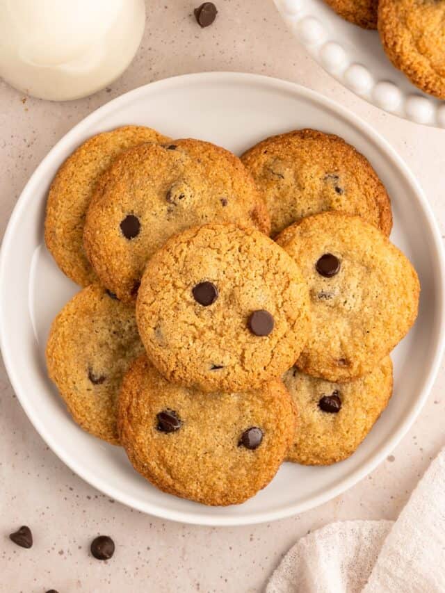 stacks of almond flour chocolate chip cookies on a white plate with a cup of milk to the side.