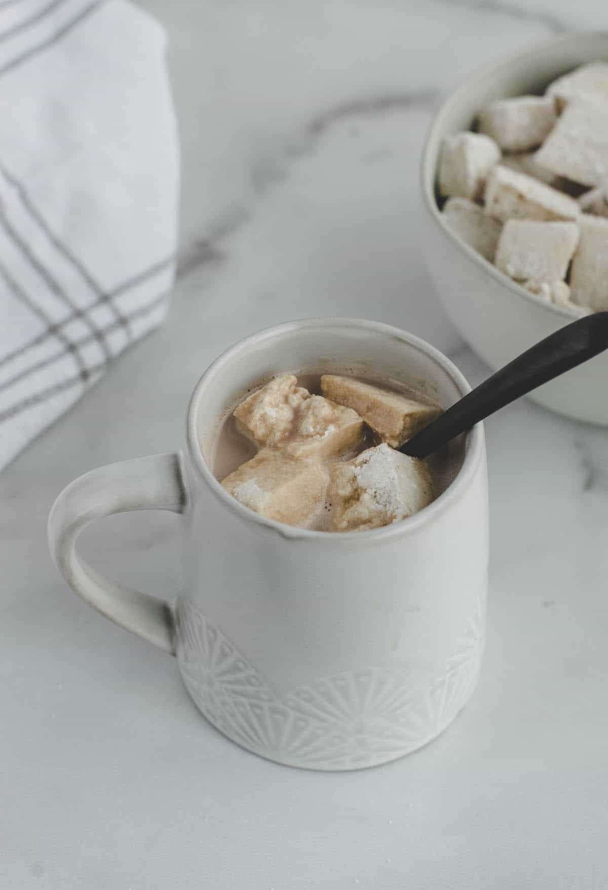 white mug with hot chocolate and healthy marshmallows and a black spoon inside of the mug. 