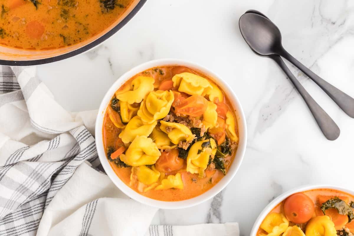 white soup bowl filled with creamy sausage tortellini soup and dutch oven to the back and two black spoons to the side of the bowl.