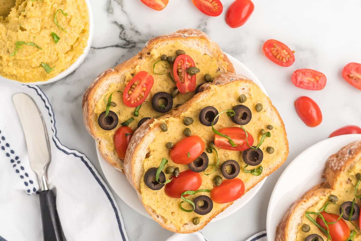 two pieces of toast with homemade hummus and topped with fresh mini tomatoes, capers, and black olives, fresh tomatoes scattered to the side and a butter knife resting on a tea towel.