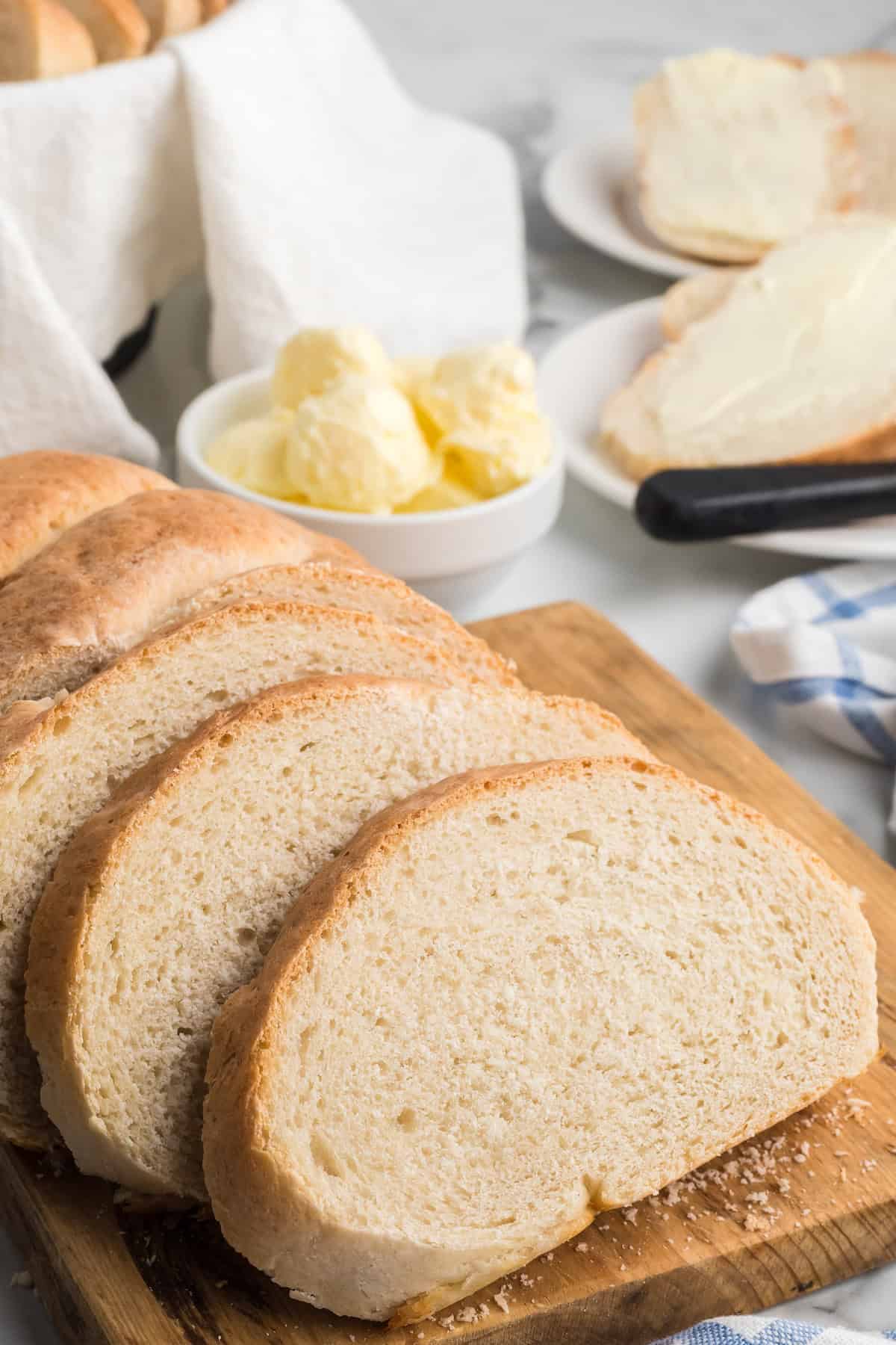 slices of italian loaf bread on a wooden cutting board with a white bowl of butter and knife to the side.