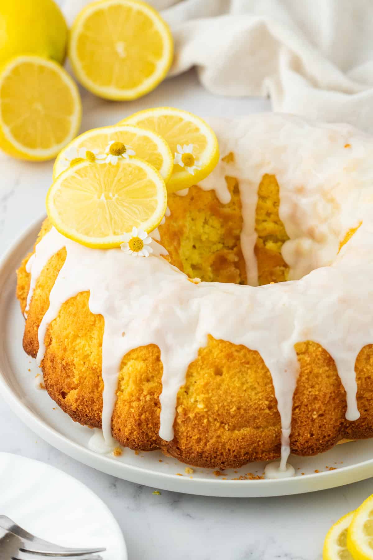 frosted bundt cake on a white plate with slices of lemon as a garnish. 