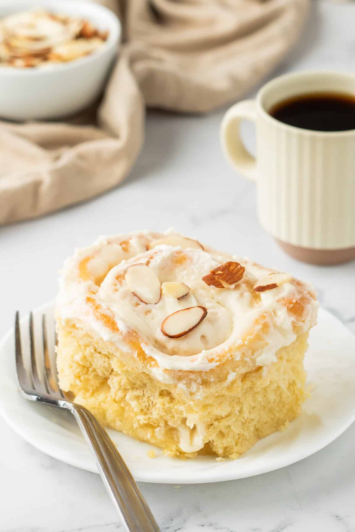 white plate with a slice of almond croissant cinnamon roll with a fork to the side. Paired with a white mug of coffee behind the plate. 