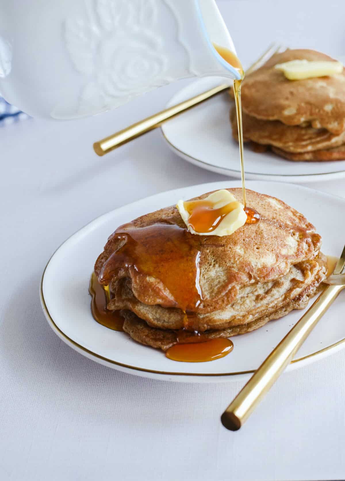 two white plates stacked with apple cinnamon sourdough pancakes and drizzling maple syrup over top. 