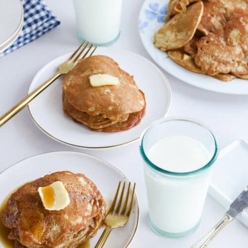 two white plates stacked with apple cinnamon sourdough pancakes and butter with drizzle of maple syrup.