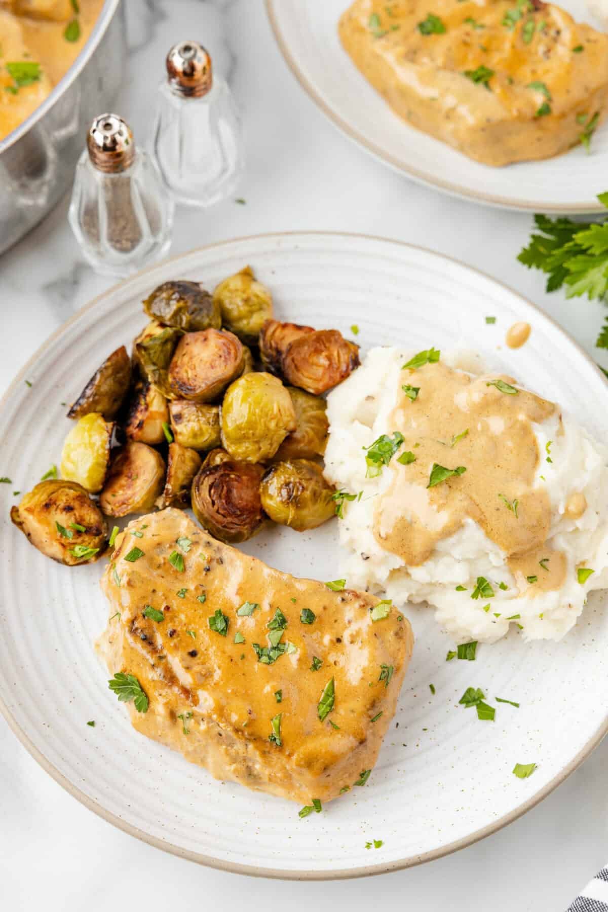 two white plates with servings of creamy pork chops, mashed potatoes and gravy, and roasted veggies with the skillet of creamy pork chops to the side. 