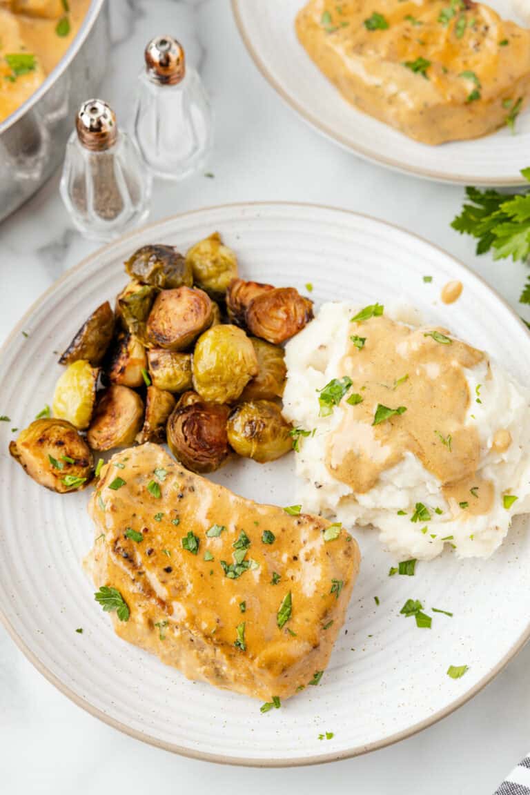 two white plates with servings of creamy pork chops, mashed potatoes and gravy, and roasted veggies with the skillet of creamy pork chops to the side.