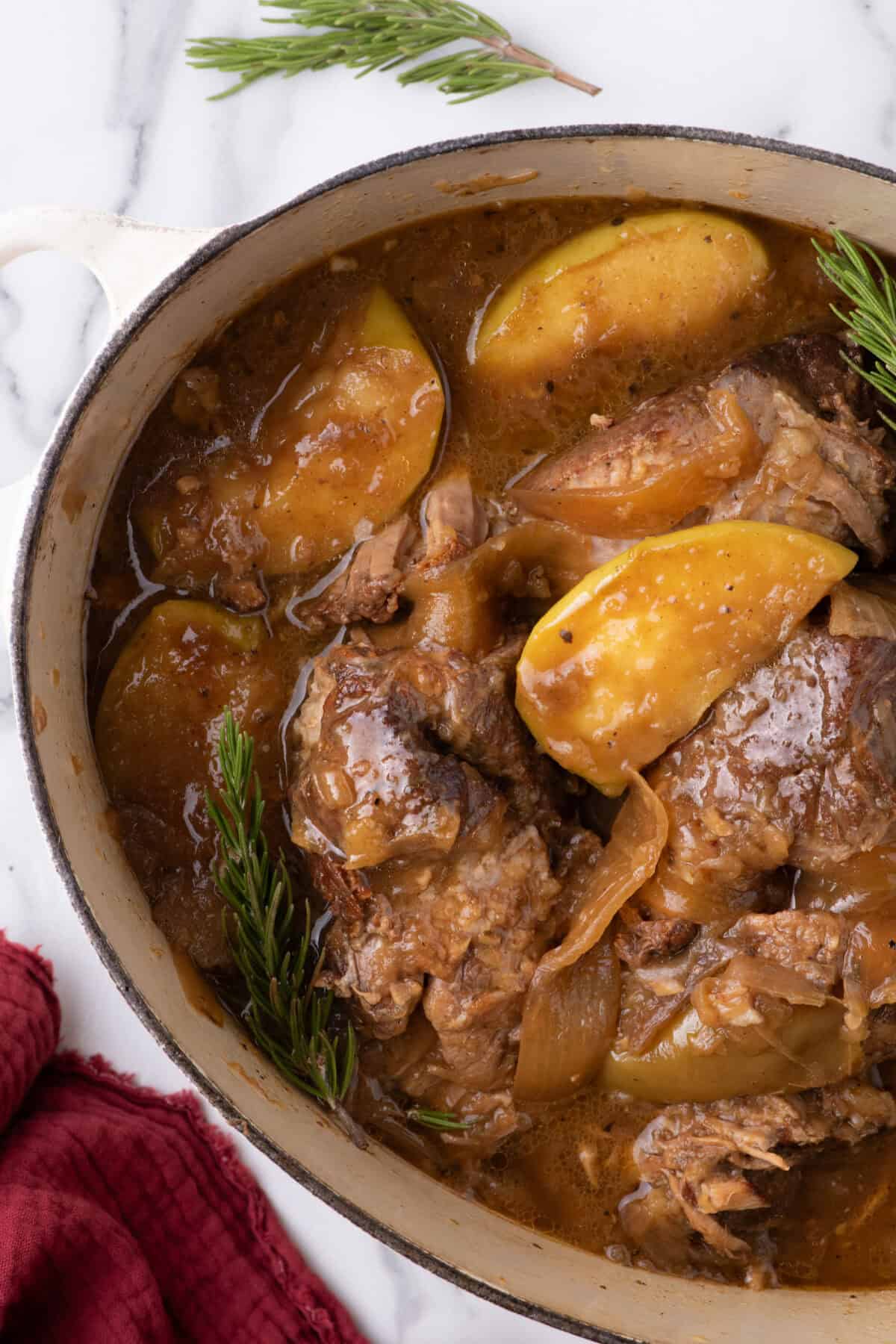 finished apple cider braised pork shoulder in a white dutch oven with cooked apples and onions over top. 