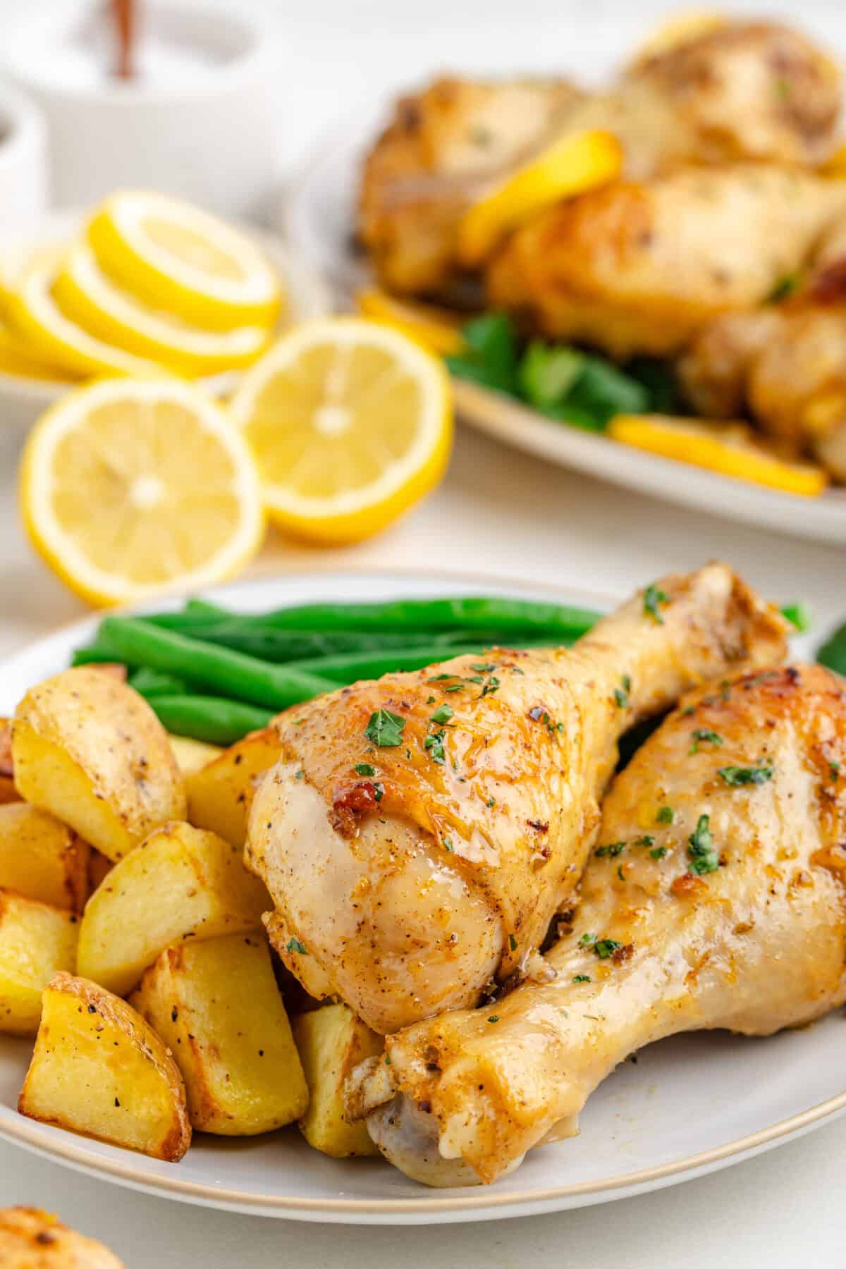 White plates stacked with baked lemon chicken drumsticks, roasted potatoes, and steamed green beans. 