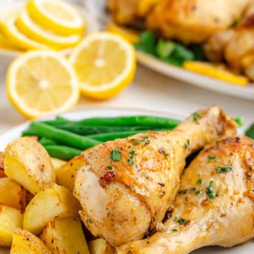 White plates stacked with baked lemon chicken drumsticks, roasted potatoes, and steamed green beans.