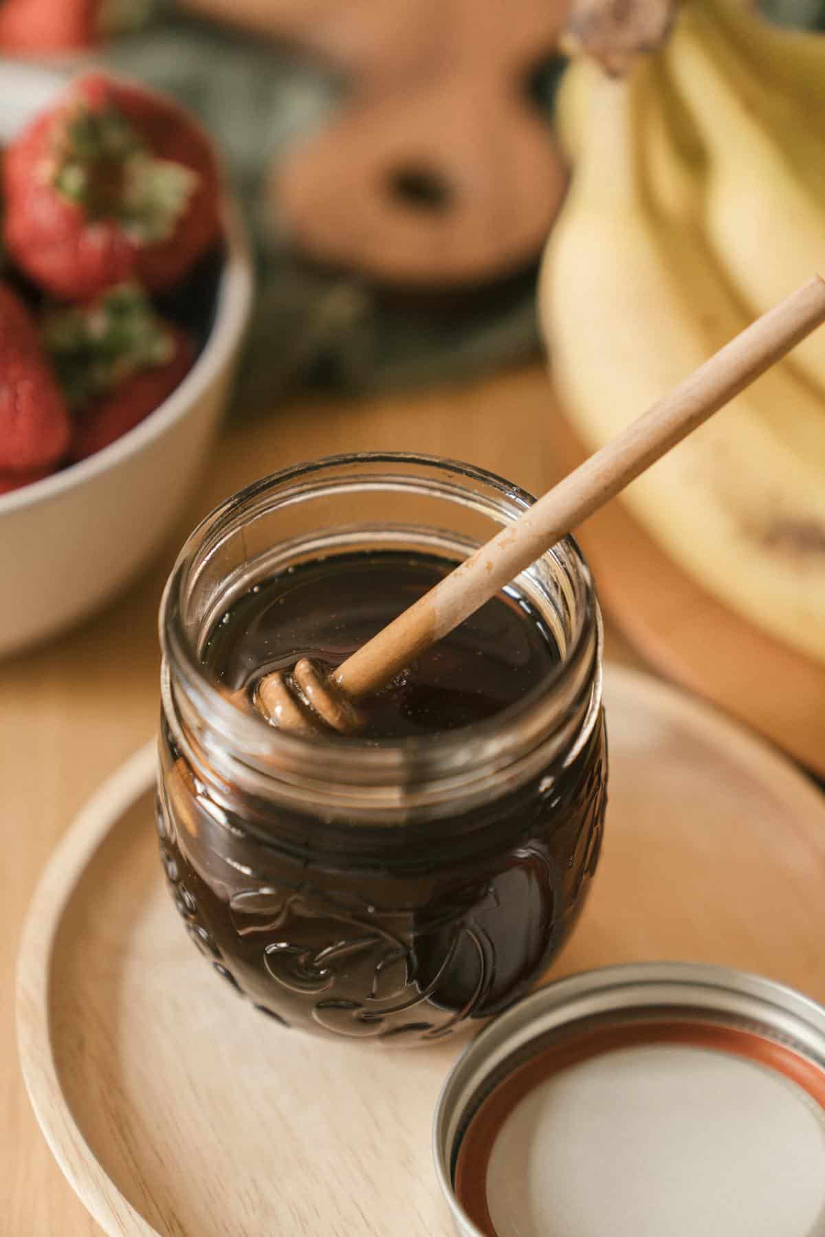 A glass jar of honey with a dipper on a wooden plate surrounded by fresh fruits.