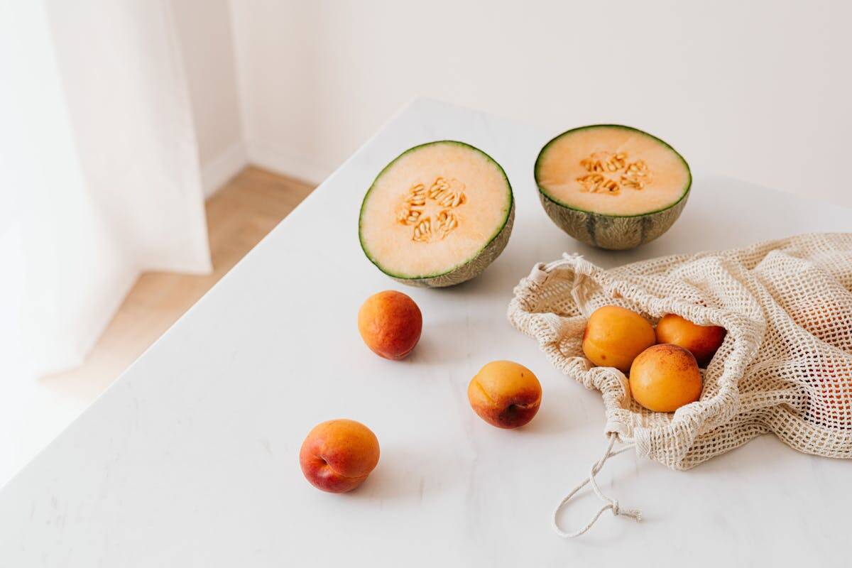 A halved melon and apricots in a mesh bag on a white marble table, captured in natural light.