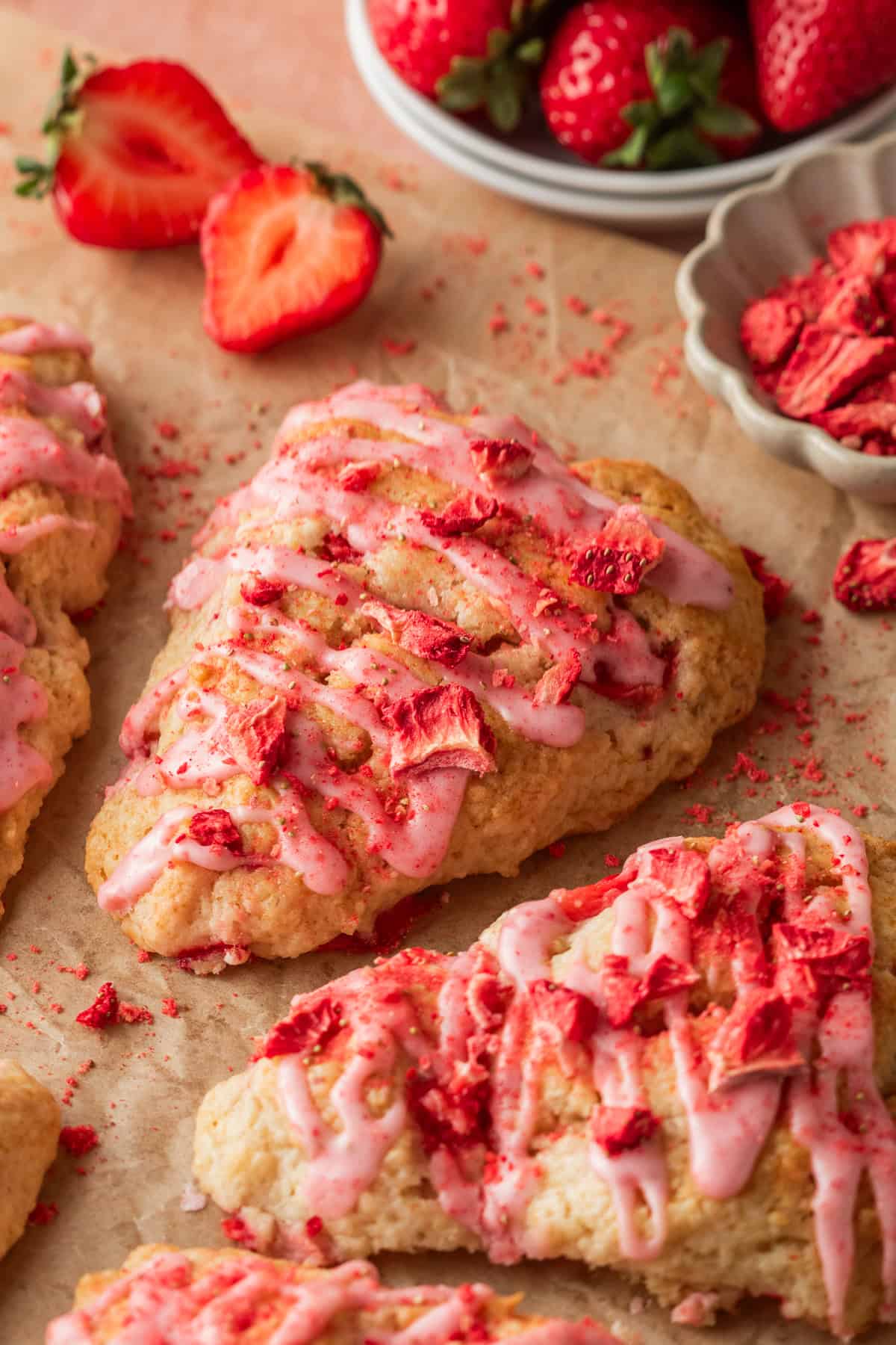 strawberry scones with strawberry glaze on parchment paper with a bowl of fresh strawberries in the background.
